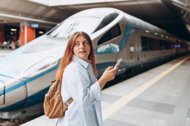Young redhhead woman waiting train on station platform with backpack and using smart phone. Railroad transport concept, Traveler