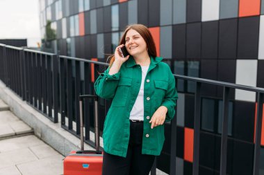 Young woman with a suitcase using smart phone at airport. Happy Redhead girl talking on the mobile phone. Urban lifestyle concept, technology banner