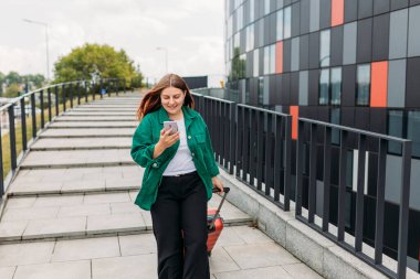 Young woman with a red suitcase using smart phone at airport. Redhead girl in international airport walking with her luggage.