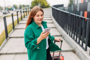 Young woman with a red suitcase using smart phone at airport. Redhead girl in international airport walking with her luggage.