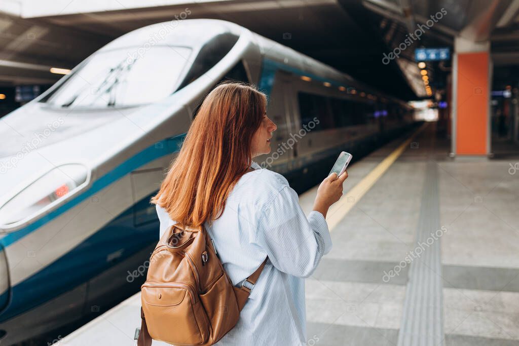 Young redhhead woman waiting train on station platform with backpack and using smart phone. Railroad transport concept, Traveler