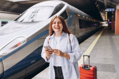Young redhhead woman waiting train with backpack and using smart phone. Railroad transport concept, Traveler.Woman with suitcase walking at railroad station platform. Travel to vacation