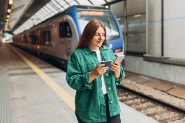 Young redhhead woman with passport waiting on station platform and using smart phone. Railroad transport concept. Travel to vacation by train