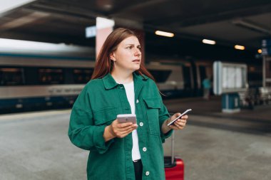 Young refugee redhhead woman waiting on station platform on background train, she lost and using smart phone. Railroad transport concept, Traveler.