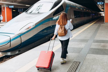 Business woman with a red suitcase using smart phone and moving on station platform for travel trip. Railroad transport and technology concept