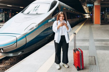 Business woman with a red suitcase using smart phone and moving on station platform for travel trip. Railroad transport and technology concept