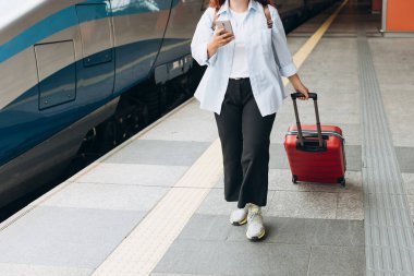 Business woman with a red suitcase using smart phone and moving on station platform for travel trip. Railroad transport and technology concept