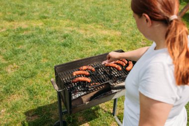 Izgara ızgarada domuz sosisi. Eti ızgara yapan genç bir kadının eli. Genç bir kadın barbekü yapıyor, genç bir kız yemek hazırlıyor.