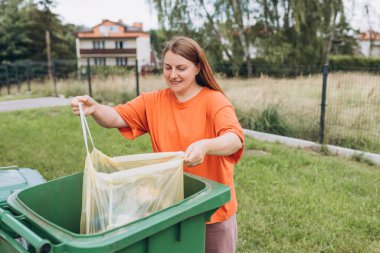 Arka bahçedeki çöpleri yeşil çöp kutusuna koyan genç bir kadın. Sarı plastik poşet tutan gülümseyen kadınlar. Çevresel koruma gezegeni kirlenmeden koruyor