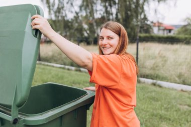 Arka bahçedeki çöpleri yeşil çöp kutusuna koyan genç bir kadın. Çevresel koruma gezegeni kirlenmeden koruyor. Plastik kirliliği sorunu.