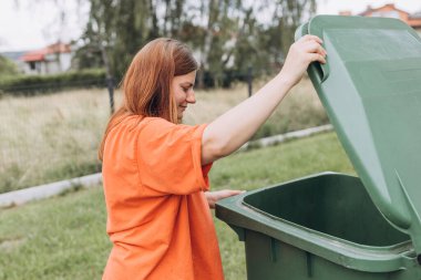 Arka bahçedeki çöpleri yeşil çöp kutusuna koyan genç bir kadın. Çevresel koruma gezegeni kirlenmeden koruyor. Plastik kirliliği sorunu.