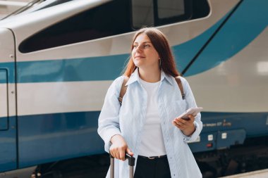 Young redhhead woman waiting train with backpack and using smart phone. Railroad transport concept, Traveler.Woman with suitcase walking at railroad station platform. Travel to vacation