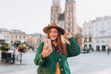 Genç bayan turist, Polonya 'nın Krakow kentindeki pazar meydanında ünlü St. Marys Bazilikası önünde selfie çekiyor.