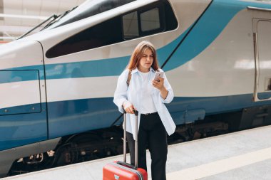 Business woman with a red suitcase using smart phone and moving on station platform for travel trip. Railroad transport and technology concept