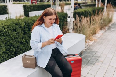 Kırmızı bavullu genç kızıl saçlı kadın şehir geçmişinde akıllı telefon kullanıyor. Modern bir bankta oturan mutlu kız. Kentsel yaşam tarzı konsepti