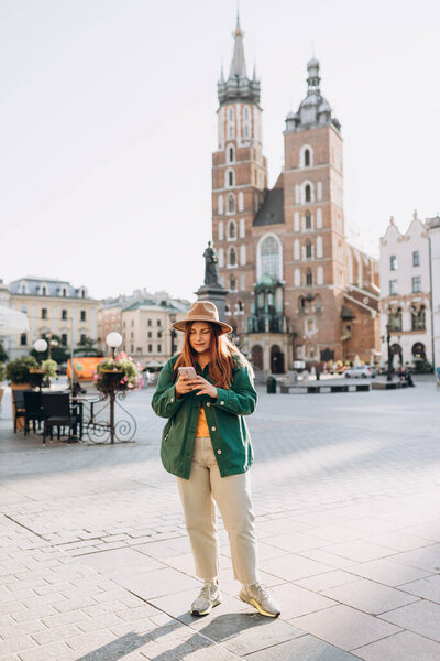 Beautiful stylish woman walking on Market Square in Krakow on autumn day and holding mobile phone. Phone Communication. Urban lifestyle concept. Check social networks
