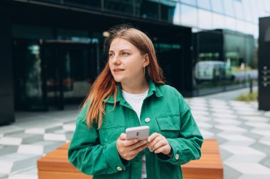 Genç mutlu iş kadını şehirde gülümsüyor ve akıllı telefon kullanıyor. Ofise giden güzel bir kadın akıllı telefonuyla internetten bilgi arıyor. İş, eğitim kavramı