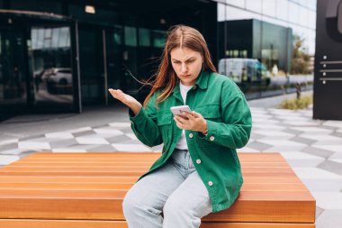 Yakından bakınca üzgün, şüpheci, mutsuz, ciddi bir kadın açık havada telefon ekranına bakıyor. Negatif insani duygu yüz ifadesi hissi. Haberler kötü. Sokakta bankta oturan kız.