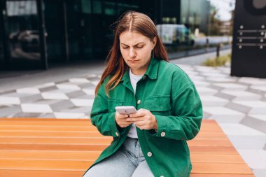 Yakından bakınca üzgün, şüpheci, mutsuz, ciddi bir kadın açık havada telefon ekranına bakıyor. Negatif insani duygu yüz ifadesi hissi. Haberler kötü. Sokakta bankta oturan kız.