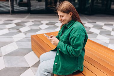 Genç mutlu kadın gülümsüyor ve şehirde akıllı telefon kullanıyor. Güzel bir kadın şehrin caddesinde bankta oturuyor ve akıllı telefonuyla internetten bilgi arıyor..
