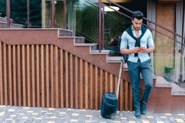 Çekici genç Arap adam elinde bavuluyla kameraya poz veriyor, telefonu tutuyor ve konuşuyor, bir ayağını diğerinden önde tutuyor ve restoranın yanındaki merdivenlere dayanıyor.