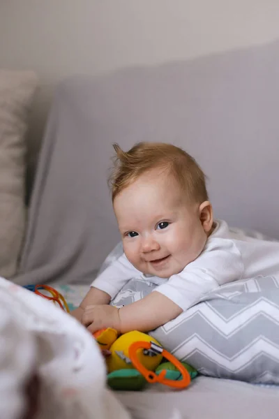 Newborn smiling baby lying on grey bedspread and playing with toys. — Stock Photo, Image