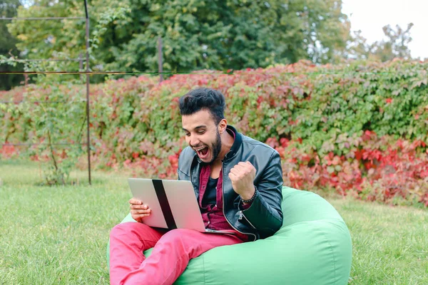 Pomyślny arabski młody biznesmen siedzi z laptopem w fotelu, gapi się na ekran laptopa, podnosi ręce, co Ręka Gesture tak w ogrodzie na zewnątrz. Obrazek Stockowy