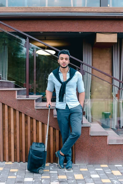 Atractivo joven árabe de pie con la maleta en la mano y posando para la cámara, poner un pie antes que el otro y se basa en las escaleras cerca del restaurante al aire libre. — Foto de Stock