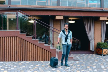 Genç adam Arap işadamı öğrenci geldiysen oraya restoran ile bavul için elinde, kamera, durur ve uzak görünüyor gülümseyen.