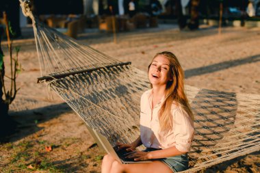 Dizüstü bilgisayar kullanarak ve hasır hamak kuma oturan genç beyaz kız.