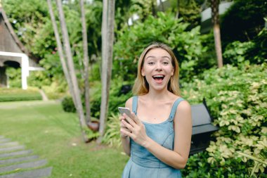 Kot sundress giyen genç güzel kadın bahçede ağaçlar, yakınındaki smartphone kullanarak.