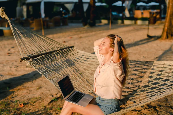 Blonde vrouwelijke persoon werken met laptop en zittend op zand in rieten hangmat. — Stockfoto