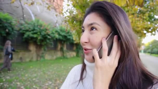 Asiática smling chica usando blanco suéter hablando por teléfono inteligente y caminando en parque . — Vídeos de Stock