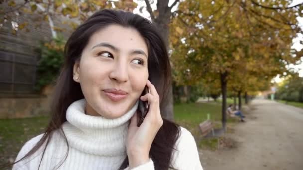 Asiática bonita chica vistiendo suéter blanco hablando por teléfono inteligente y caminando en parque . — Vídeos de Stock