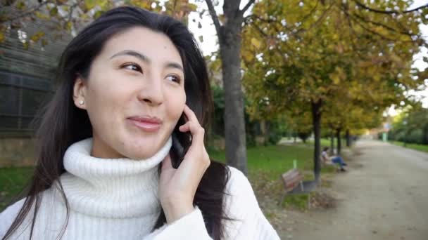 Cámara lenta primer plano asiático smling chica usando blanco suéter hablando por smartphone y caminando en parque. — Vídeo de stock