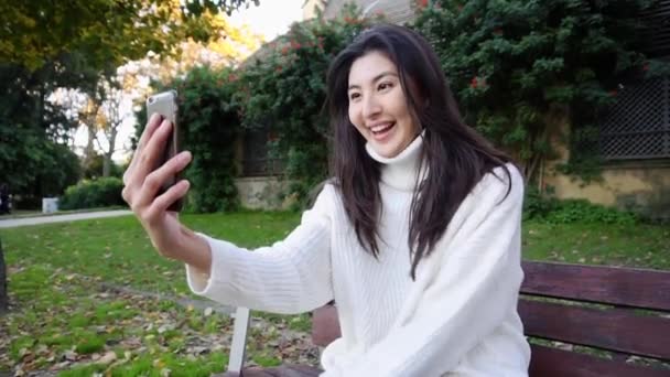 Chica japonesa sentada en el banco y haciendo videollamada por teléfono inteligente . — Vídeo de stock