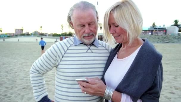 Birlikte bir cep telefonuyla konuşan bir sahilde bir masada oturan mutlu kıdemli adam ve kadın çift — Stok video