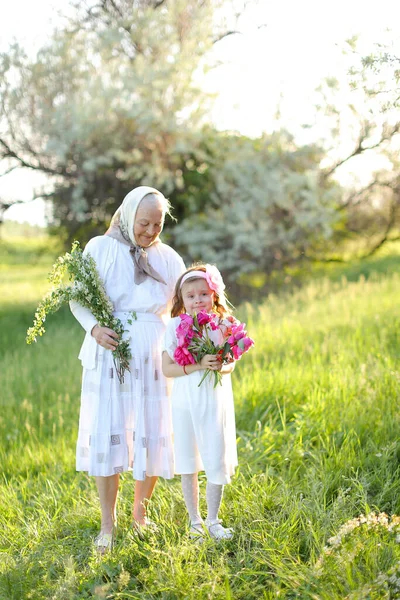 Velha avó caucasiana de pé com pequena neta e mantendo flores. — Fotografia de Stock