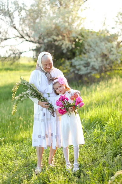 Old grandmother standing with little granddaughter and keeping flowers. — Stock Photo, Image