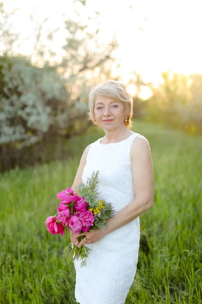Caucasian blonde woman standing with flowers in park, wearing white dress and hat. — Stock Photo, Image