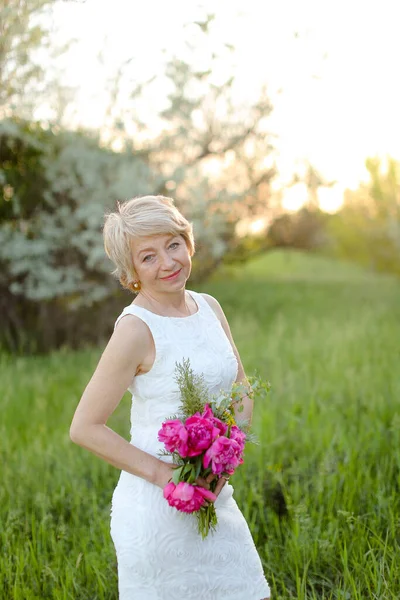 Caucasian european woman standing with flowers in park, wearing white dress and hat. — Stock Photo, Image