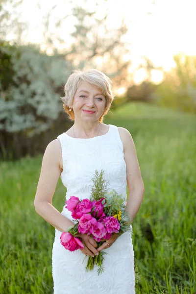 Caucasian smiling woman standing with flowers in park, wearing white dress and hat. — Stock Photo, Image