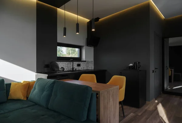 View of modern studio apartment with green sofa and yellow dining chairs in dark kitchen with copy space