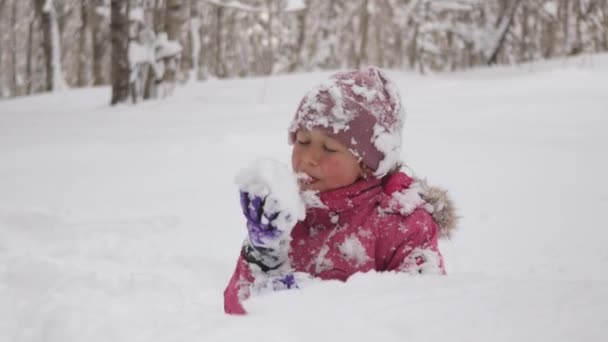 小さなかわいい子供の女の子は森の中の雪のドリフトに座って食欲をもって雪を食べている — ストック動画