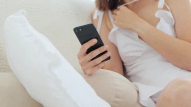 Beyaz pijamalı genç bir kadın sabahları kanepeye otururken akıllı telefon kullanıyor, 4K yakın plan.