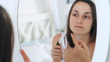 Yüz temeli ya da nemlendirici krem sürerken aynaya bakan 30 'ların beyaz kadın portresi. Cilt bakımı ve kozmetik konsepti, yakın plan 4k 