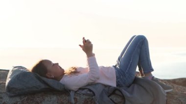 Kız deniz kıyısında yatıyor, taşların üzerinde, akıllı telefonuyla oyunlar oynuyor, 4K video.