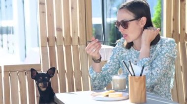 Çekici kadın küçük köpeğiyle kafede oturur ve evcil hayvanıyla kahvaltısını paylaşır. 4K görüntüler.
