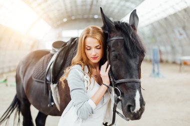 beautiful blond professional female jockey standing near horse. woman horse rider is preparing to equitation. girl and horse. equestrian sport concept. riding vacation.friendship with horse.copy space