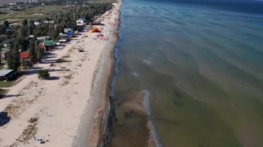 Yazın sahilde güzel bir uçuş. İnsanlar deniz kenarında dinleniyor. Turistler için evler. Plaj şemsiyeleri, gezintiler, denizde yüzen insanlar..